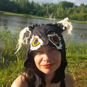 May include: A woman wearing a black felt owl hat with white fur trim and orange and yellow felt eyes. The hat has a hood and covers her head and ears. She is standing in front of a body of water with green grass and trees in the background.