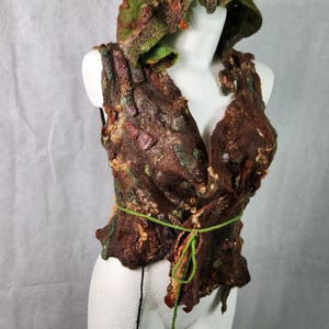 Puede incluir: Un chaleco artesanal de fieltro marrón con capucha adjunta. El chaleco presenta un cuello en V y una cinta verde en la cintura. La capucha y el chaleco están adornados con detalles verdes, naranjas y beige, creando una apariencia texturizada y orgánica.