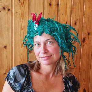 Puede incluir: Una mujer con un sombrero azul turquesa con un adorno de flor roja y blanca. El sombrero tiene largos hilos de lana que cuelgan. Lleva una blusa de estampado de camuflaje.