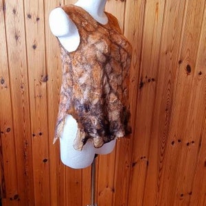May include: A sleeveless top made of felted wool with a brown and beige marbled pattern. The top is displayed on a white mannequin on a tripod stand against a wood panel background.