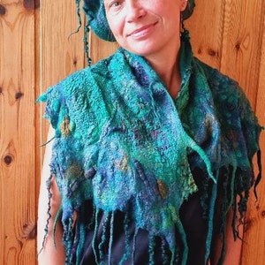 May include: A woman wearing a teal blue and green felted wool scarf with long fringe. The scarf has a mottled pattern and is draped around her neck. She is also wearing a teal blue and green felted wool hat.