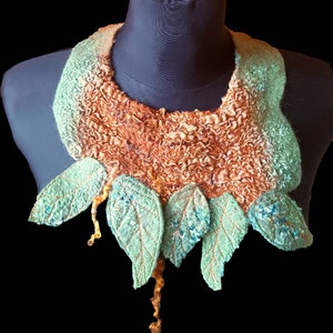 May include: A felted wool necklace with a brown and green color gradient. The necklace features a ruffled collar with a row of green leaves attached to the bottom.
