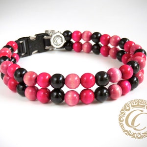 Può includere: Un collare per animali domestici con perline di legno nere e rosa alternate. Il collare ha una fibbia in plastica nera e un ciondolo color argento. Le perline sono disposte in due file parallele.