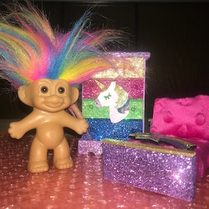 May include: A vintage troll doll with rainbow hair stands next to a miniature glittery bedroom set. The set includes a cabinet with a unicorn design and a pink armchair. The furniture is decorated with glitter.