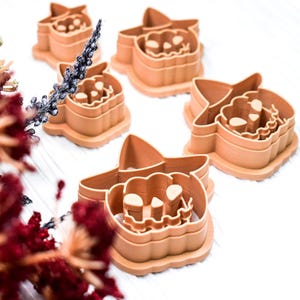 May include: A collection of tan plastic cookie cutters in the shape of a jack-o'-lantern wearing a witch's hat. The cutters are arranged on a white surface, with dried flowers in the foreground.