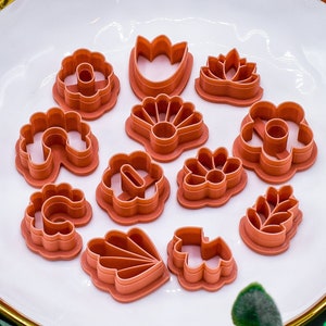 May include: A set of 12 terracotta-colored cookie cutters shaped like flowers, leaves, and other floral designs. The cutters are arranged on a white plate with a gold rim.