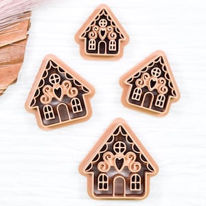 May include: Four brown gingerbread house cookie cutters on a white wooden surface. The cookie cutters are detailed with hearts and swirls.