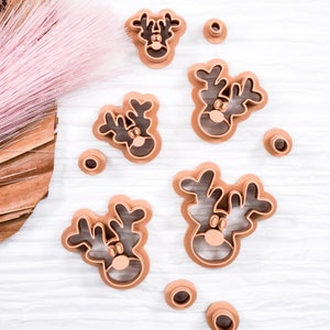 May include: A set of six brown plastic reindeer cookie cutters on a white wooden surface. The cutters are arranged in a circle with a few small brown circles scattered around them.