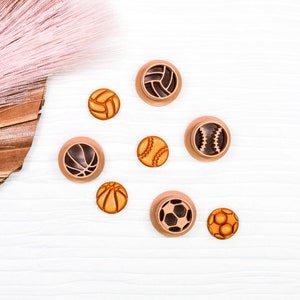 May include: A collection of eight small, round, wooden buttons with different sports ball designs. The buttons are arranged in a semi-circle on a white wooden surface. The buttons are brown and orange with black details.