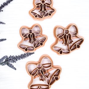 May include: Four brown Christmas bell cookie cutters with bow details. The cutters are arranged on a white surface, with decorative sprigs in the upper left corner. The bell shapes have a detailed design, perfect for holiday baking.
