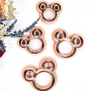 May include: Four copper-colored cookie cutters in the shape of a mouse head with large round ears. The cutters are arranged on a white surface, with dried flowers in the upper left corner.