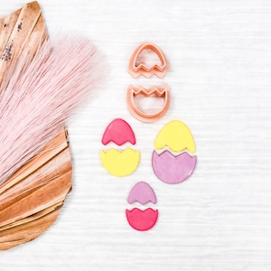 May include: A flat lay of pink, yellow, and purple fondant Easter eggs with two pink plastic cookie cutters shaped like eggs. The eggs are on a white wooden surface with a dried palm leaf and pink feathers in the background.