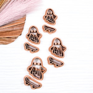 May include: Four brown plastic cookie cutters shaped like ghosts riding skateboards. The cutters are arranged in a vertical line on a white wooden surface.