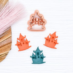 May include: Three cookie cutters in the shape of a haunted house. One is a light brown plastic cutter, and the other two are made of colored fondant. One is orange and the other is teal.