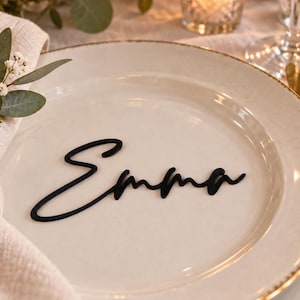 May include: A black script name tag reading Emma placed on a cream-colored ceramic dinner plate with a gold-trimmed rim. The plate is set with a beige linen napkin and a small sprig of white baby's breath flowers and green eucalyptus leaves.