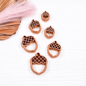 May include: Five brown plastic acorn-shaped cookie cutters of varying sizes. The cutters are arranged on a white wooden surface.