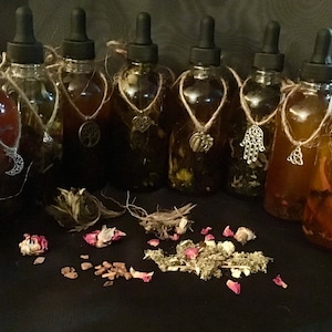 May include: Ten glass bottles with dropper tops filled with a dark liquid. Each bottle has a silver charm hanging from a twine necklace around the neck of the bottle. The bottles are arranged in a row on a black surface.
