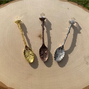 Puede incluir: Tres cucharas decorativas con mangos texturizados en forma de rama y remates de cristal transparente. Las cucharas presentan patrones florales en relieve en la parte cóncava y tienen acabados en tonos oro, bronce oscuro y plata pulida.