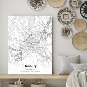 May include: A black and white map print of Danbury, Connecticut, with coordinates. The map is displayed on a white canvas, with a neutral-toned bench and woven baskets below. Decorative wall hangings and pillows add to the decor.