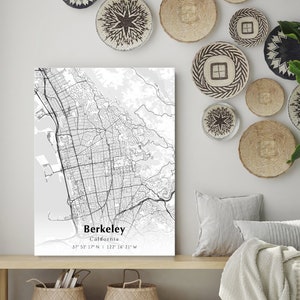 May include: A black and white map print of Berkeley, California with the city's name and coordinates printed below the map. The print is hanging on a white wall with woven wall decor.