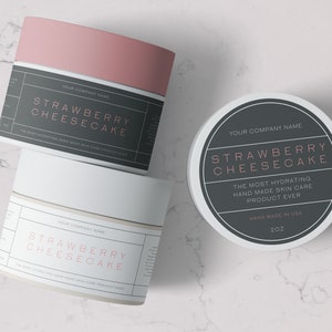 May include: Three white jars with black labels and pink text. The labels read "Strawberry Cheesecake" and "The Most Hydrating Hand Made Skin Care Product Ever". The middle jar has a black lid, the other two have pink lids. The jars are on a white marble surface.
