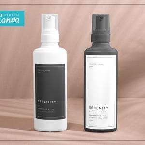 Op de afbeelding: Twee witte flessen met zwarte etiketten en zwarte pompjes. De etiketten lezen "Serenity" en "Kamille & Lelie" met de tekst "Aromatherapie-kamerspray" eronder.