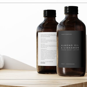 May include: Two brown glass bottles of almond oil and cinnamon vegan body lotion. The bottles have black labels with white text that reads "Almond Oil & Cinnamon Vegan Body Lotion" and "Your Company". The bottles are sitting on a wooden surface.