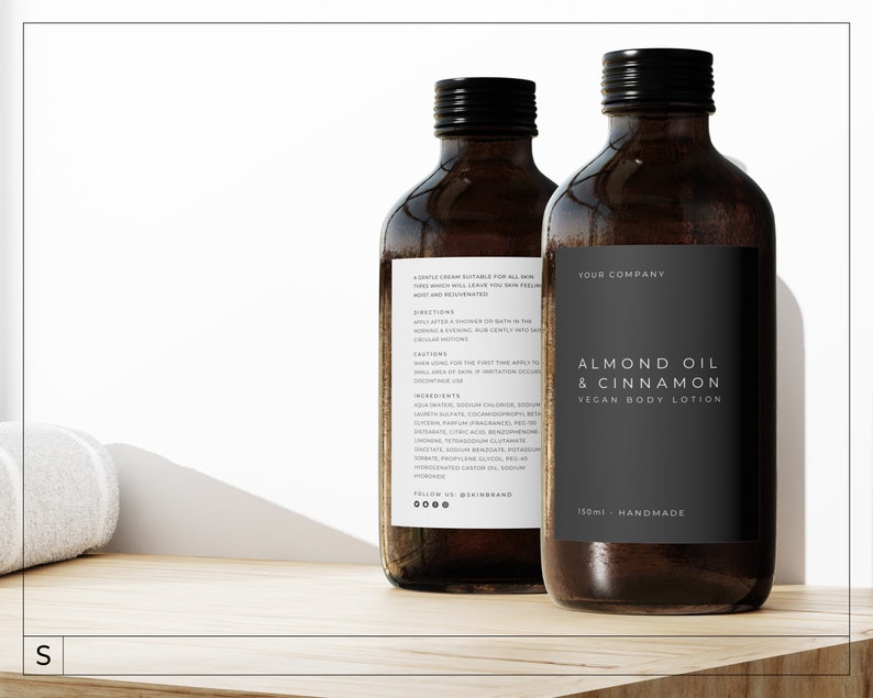 May include: Two brown glass bottles of almond oil and cinnamon vegan body lotion. The bottles have black labels with white text that reads "Almond Oil & Cinnamon Vegan Body Lotion" and "Your Company". The bottles are sitting on a wooden surface.