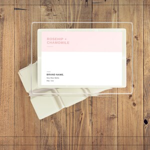 May include: A clear plastic container with a white label that reads "ROSEHIP + CHAMOMILE" in pink text. The label also includes the text "BRAND NAME. Soy Wax Melts 60g / 2 oz."