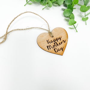May include: A wooden heart-shaped tag with a twine string attached. The tag is light brown and has the text "Happy Mother's Day" engraved on it.
