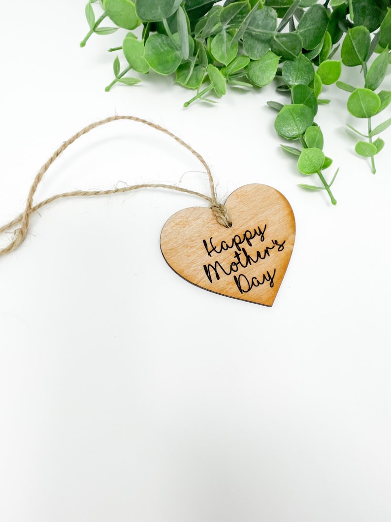 May include: A wooden heart-shaped tag with a twine string attached. The tag is light brown and has the text "Happy Mother's Day" engraved on it.