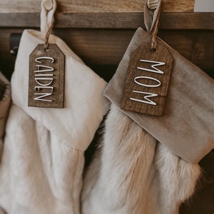 May include: Two Christmas stockings with personalized wooden tags. The stocking on the left is white with a tag that says "Caiden". The stocking on the right is brown with a tag that says "Mom".