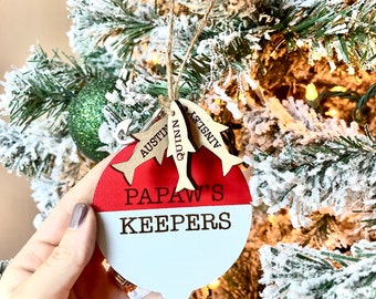 May include: A red and white wooden ornament shaped like a fishing bobber with the text "Papaw's Keepers" and three small wooden fish with the names "Ainsley", "Quinn", and "Austin" hanging from it.