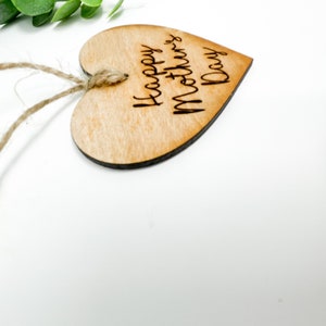May include: A wooden heart-shaped ornament with a twine string attached. The ornament is engraved with the text "Happy Mother's Day".