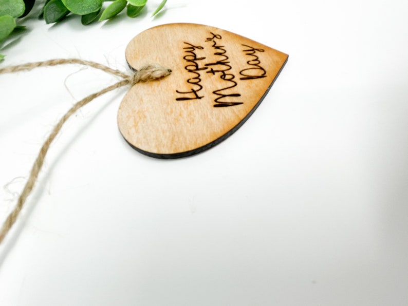 May include: A wooden heart-shaped ornament with a twine string attached. The ornament is engraved with the text "Happy Mother's Day".