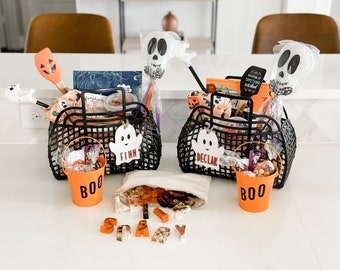 May include: Two black plastic baskets filled with Halloween items, including skull-shaped wands, orange spatulas, and books. Each basket has a white ghost tag labeled Finn or Declan. Two orange buckets labeled BOO and a canvas pouch with letter-shaped resin pieces.