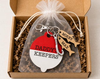 May include: A keychain set featuring a round red and white fishing bobber charm with the text DADDY'S KEEPERS, a silver metal fish hook, and two small wooden fish-shaped charms engraved with the names MIKEY and MAX, all inside a sheer white bag.
