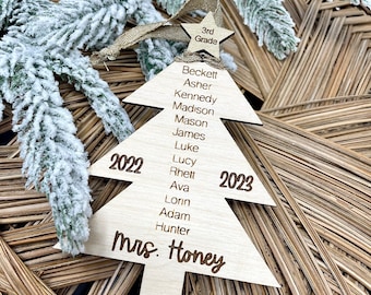 Może przedstawiać: Drewniana ozdoba choinkowa w kształcie choinki z imionami uczniów i nauczycielki, "Mrs. Honey", oraz datami "2022" i "2023". Ozdoba ma również gwiazdę na szczycie z napisem "3rd Grade".