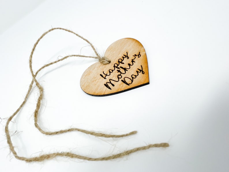 May include: A wooden heart-shaped tag with the text "Happy Mother's Day" engraved on it. The tag is attached to a piece of twine.