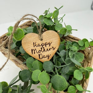 May include: A wooden heart-shaped ornament with the text "Happy Mother's Day" engraved on it. The ornament is hanging from a twine string and is surrounded by green foliage.