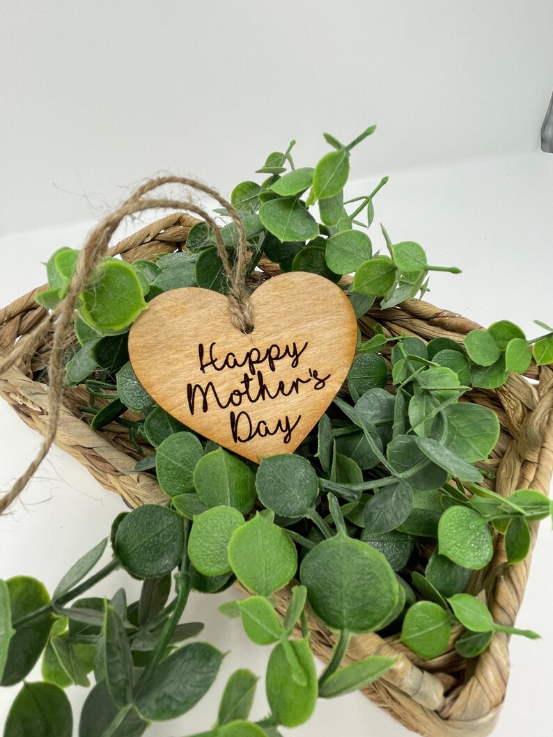 May include: A wooden heart-shaped ornament with the text "Happy Mother's Day" engraved on it. The ornament is hanging from a twine string and is surrounded by green foliage.
