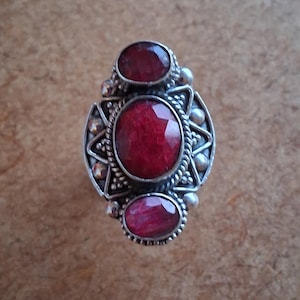 May include: Silver ring featuring three red gemstones. The central, faceted stone is surrounded by a decorative silver setting with star and bead details. The ring has an ornate, vintage design, suitable for a special occasion.