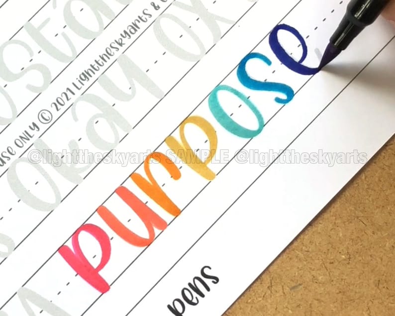 May include: A close-up of a white paper with the word "Purpose" written in colorful calligraphy. The word is written in shades of red, orange, yellow, teal, and blue. The image also includes the text "@lighttheskyarts SAMPLE @lighttheskyarts".