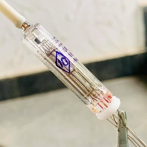 May include: A clear glass vacuum tube with internal filaments and a white ceramic base. A thin wire extends from the top, and the tube is held by a metal clamp. The tube has blue markings.