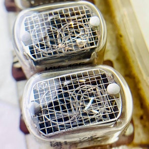 Puede incluir: Primer plano de tres tubos Nixie de vidrio transparente con un patrón de rejilla y un punto blanco en la parte superior. Los tubos están montados en una placa de metal.