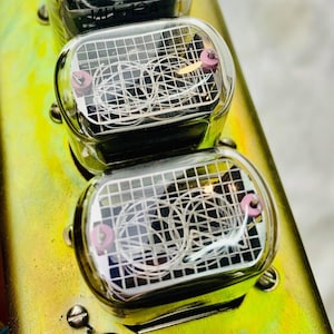 May include: Close-up of a vintage electronic device with four clear glass tubes. Each tube has a grid pattern and a small pink dot on the top. The device is mounted on a yellow metal plate.