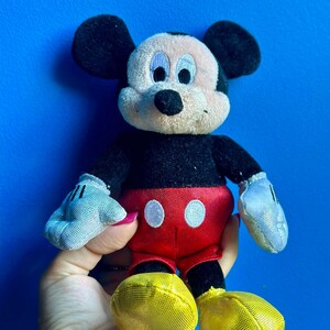 May include: A plush Mickey Mouse toy with black fur, large ears, and a red and white outfit. The toy has a yellow mesh fabric on the shoes.