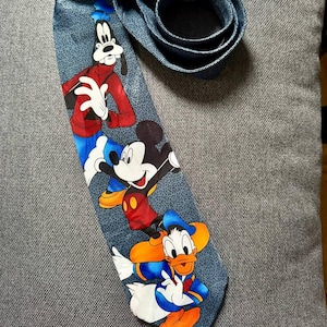 May include: A blue necktie featuring cartoon characters Goofy, Mickey Mouse, and Donald Duck. The tie has a textured pattern and the characters are in their classic outfits. The tie is rolled up at the top.