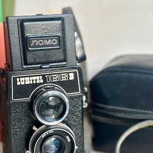 May include: A black Lubitel 166B camera with a LOMO logo on the top. The camera has a lens with a 4/135mm inscription and a black leather strap.