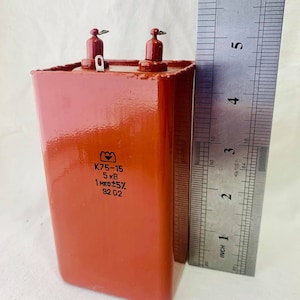 Puede incluir: Un condensador rectangular rojo con dos terminales metálicos. El condensador está etiquetado como "K75-15 5 kB 1 мкФ±5% 92 02". El condensador se mide con una regla, mostrando que tiene aproximadamente 7,6 cm de altura.